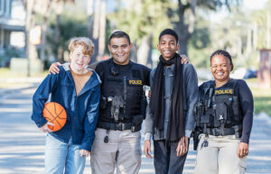 Community policing - two police officers standing with two youths in a public park. One officer, a young Hispanic man, is standing between the two boys, an African-American 14 year old teenager, and a 12 year old boy holding a basketball. The other police officer is an African-American woman. They are smiling at the camera.